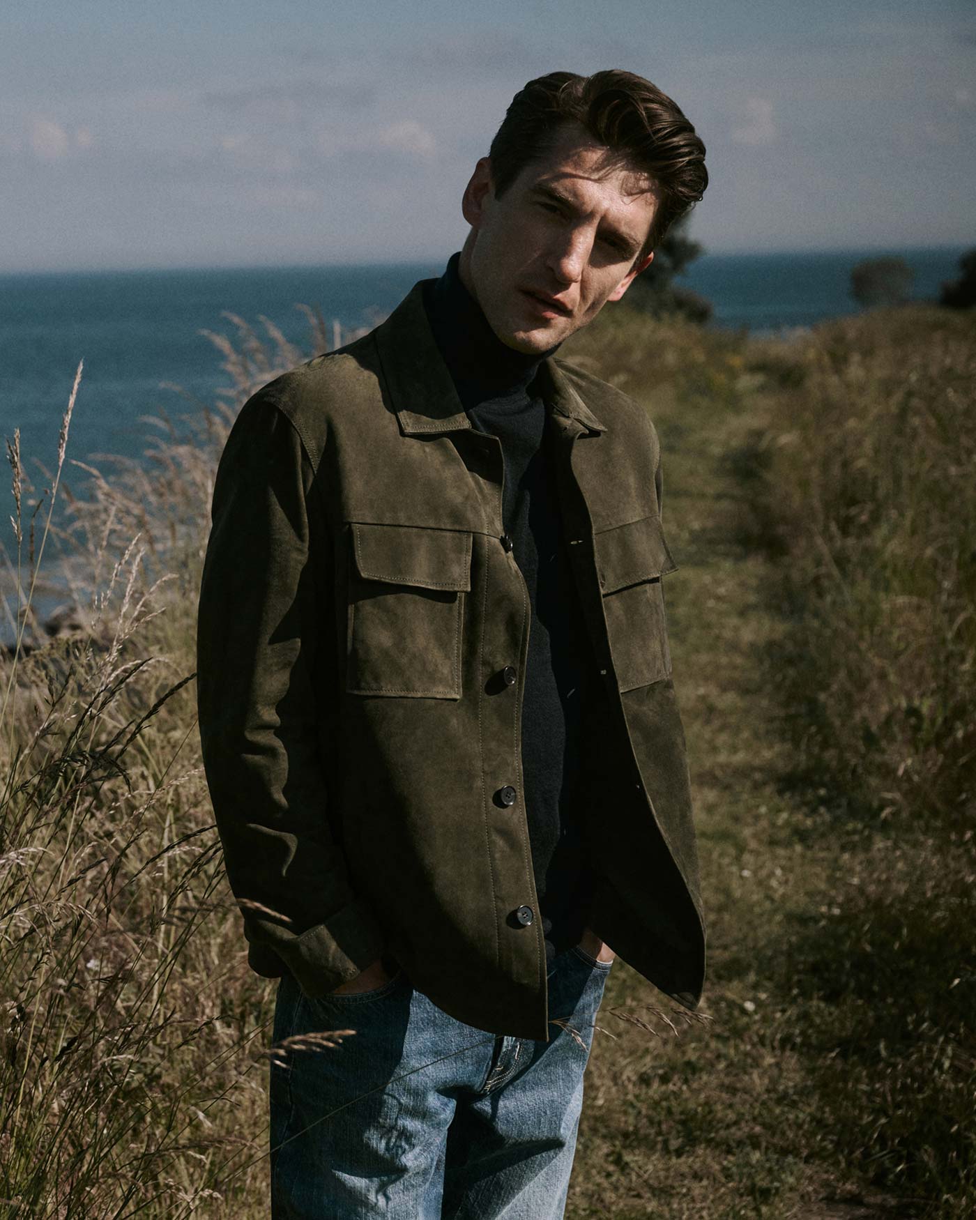 Mann mit dunkelgrüner Jacke und Jeans steht in einer Wiese, im Hintergrund Meer und bewölkter Himmel.