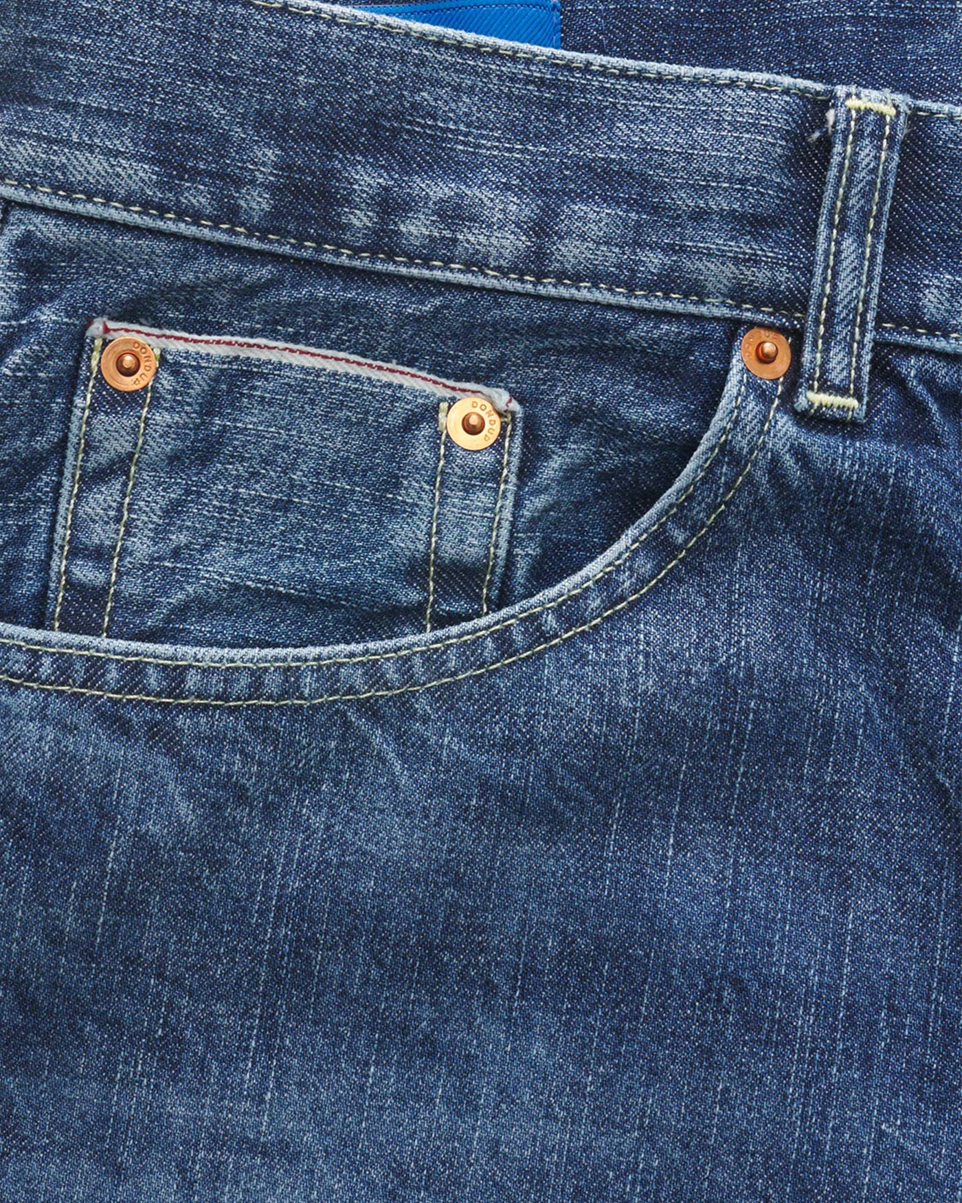 Jeansstoff in Nahaufnahme mit kleiner Münztasche, Kupfernieten, sichtbaren Nähten und einem blauen Etikett im Bund.